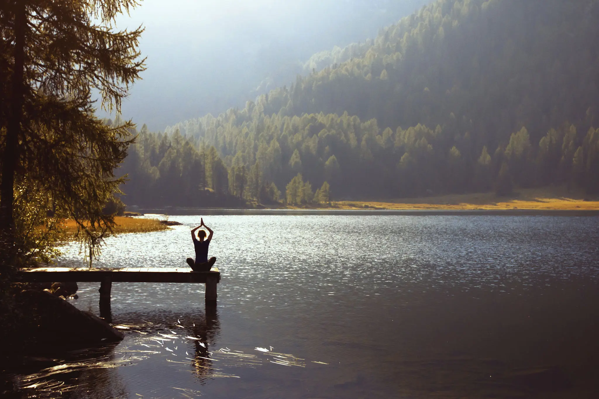 Person meditating on dock by serene lake.